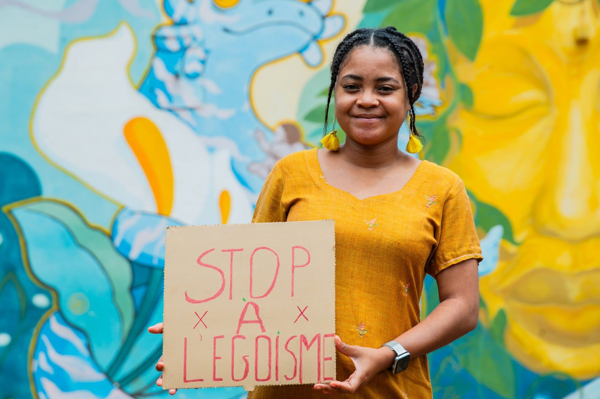Francia tenant une pancarte « Stop à l’égoïsme » devant une fresque murale