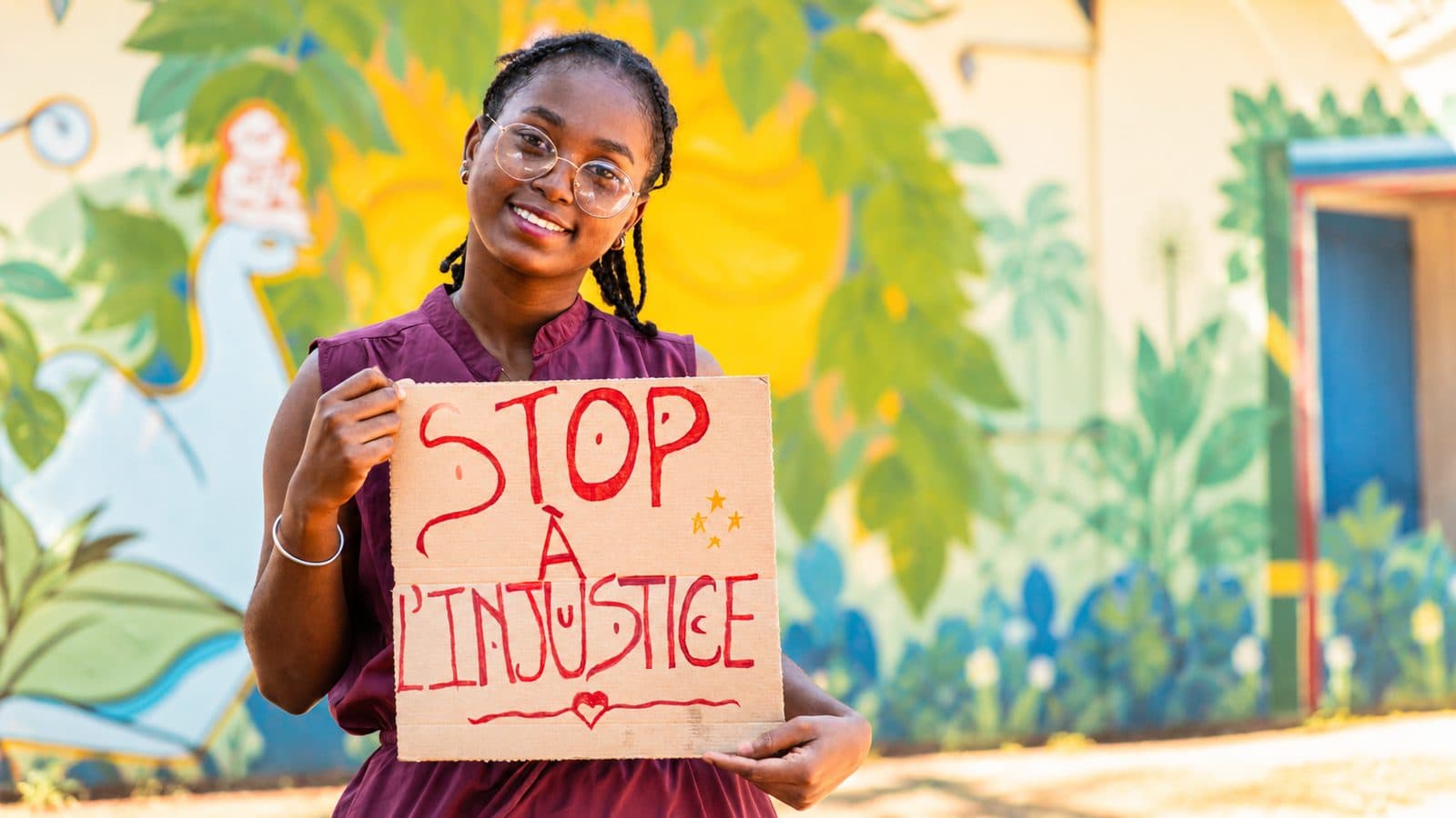 Une femme de l’association tenant une pancarte « Stop à l’injustice » devant une fresque murale